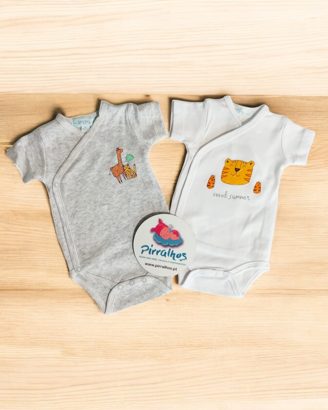 Dois bodys de bebé cinzento com girafa e branco com tigre, em fundo de madeira