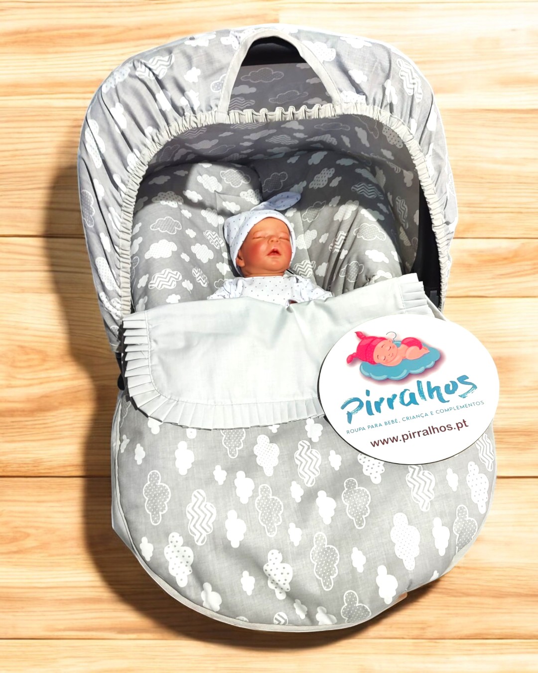 Saco-cama para bebé cinzento com padrão de nuvens brancas, boneco bebê dentro e etiqueta Pirralhos