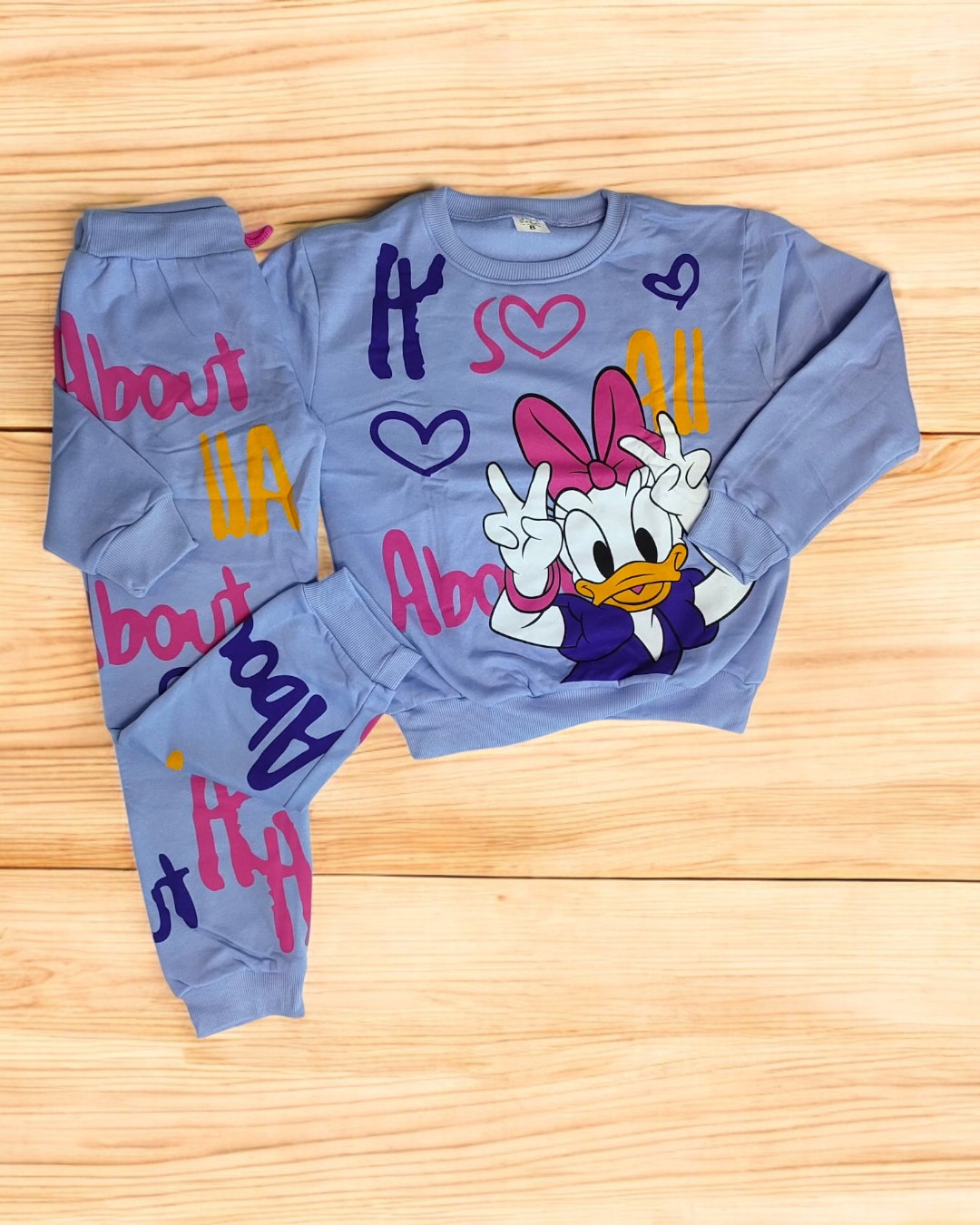 Conjunto de roupa infantil azul com estampa da Margarida e textos coloridos, sobre fundo de madeira clara.