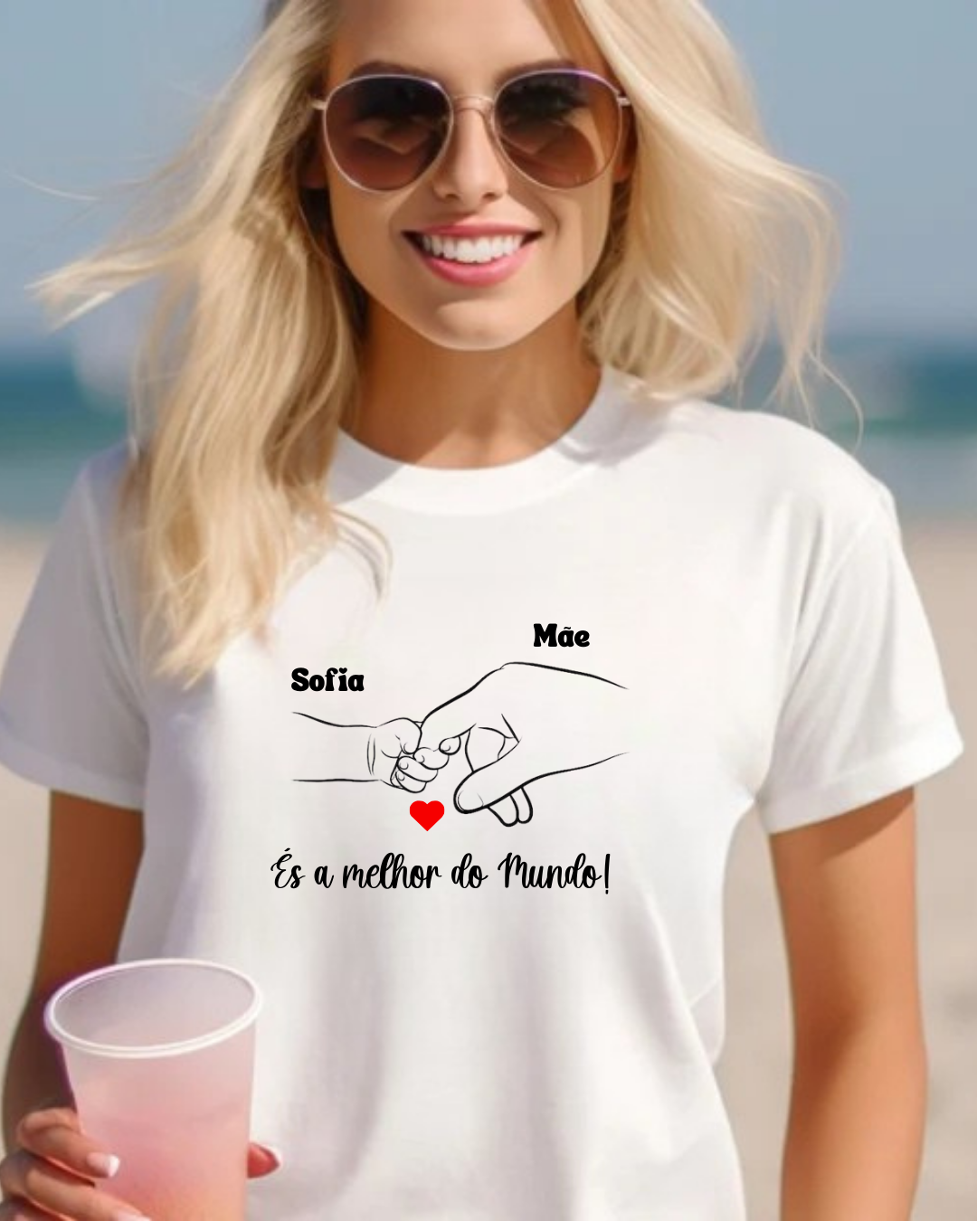 Mulher loira de óculos escuros a usar t-shirt branca com design de mãos e texto em português, segurando copo cor-de-rosa na praia.