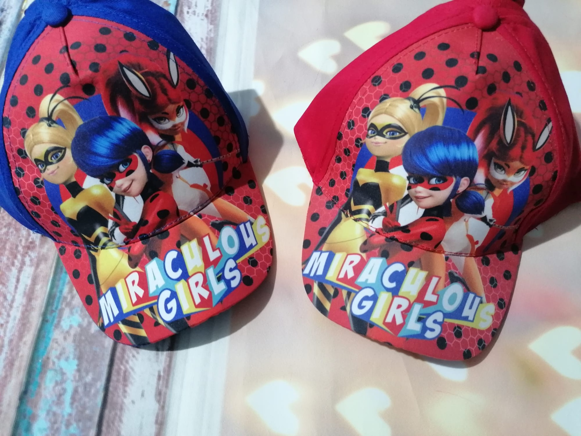 Dois bonés vermelhos e azul com estampas das personagem Miraculous Girls e texto colorido na aba