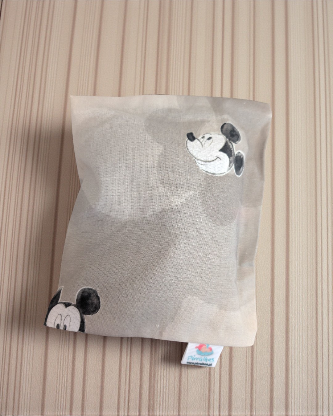 Almofada cinza clara com estampas do rosto do Mickey Mouse em preto e branco sobre superfície de madeira clara.