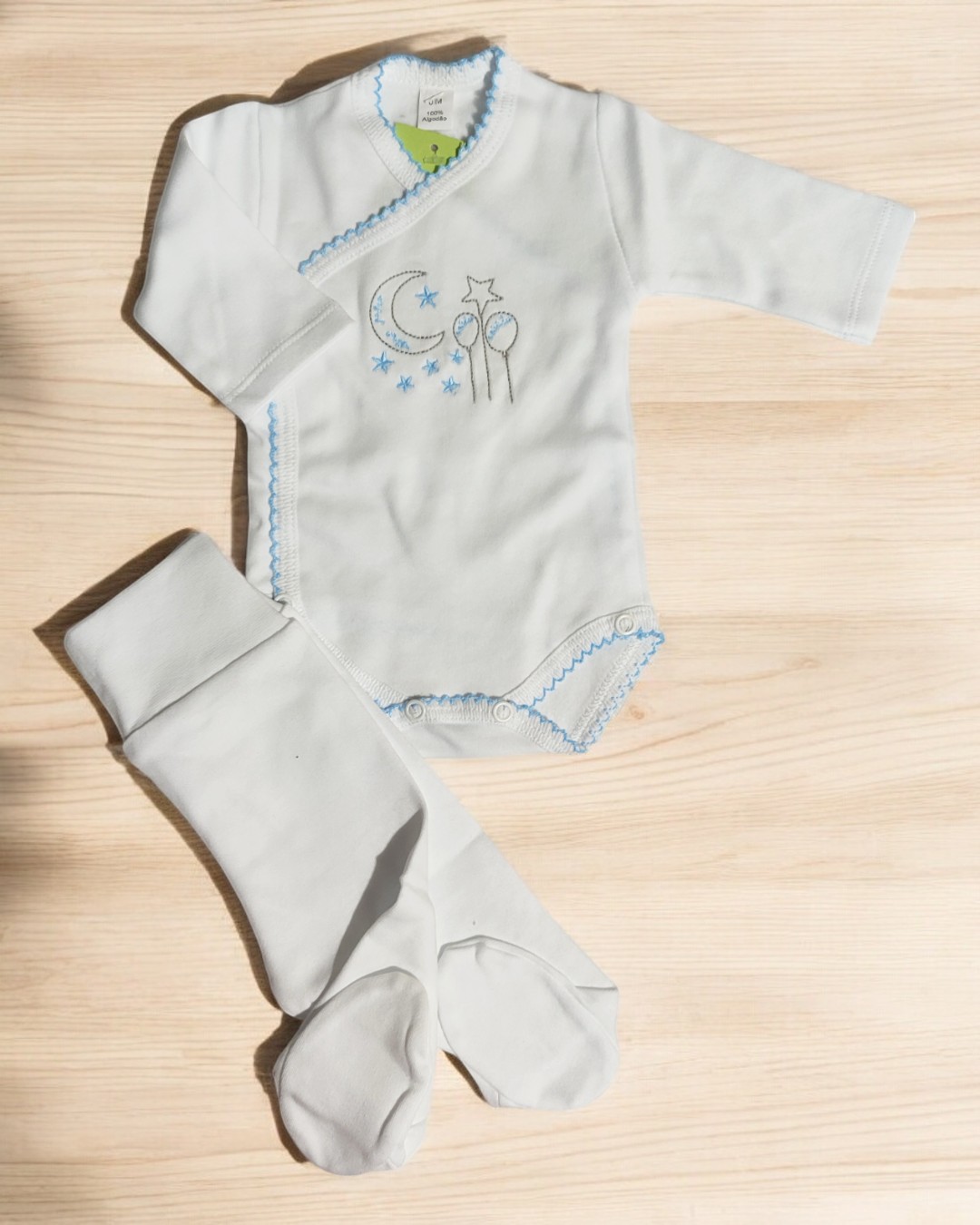Conjunto de bebé branco com bordados azuis e meia-calça branca sobre fundo de madeira clara
