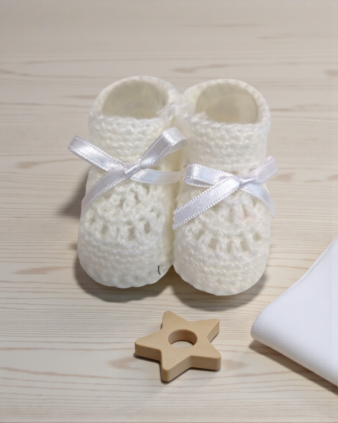 Pantufas brancas de bebé em malha grossa com laços brancos sobre mesa clara, estrela de madeira e tecido branco.