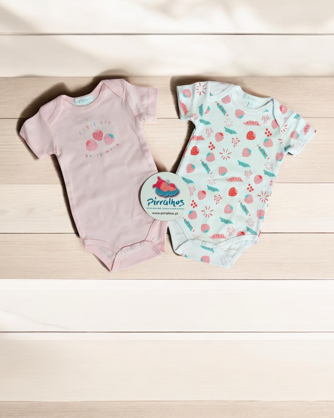 Dois bodies para bebé, um cor-de-rosa e um branco com padrão de morangos, sobre madeira clara com etiqueta 'Pirralhos'.