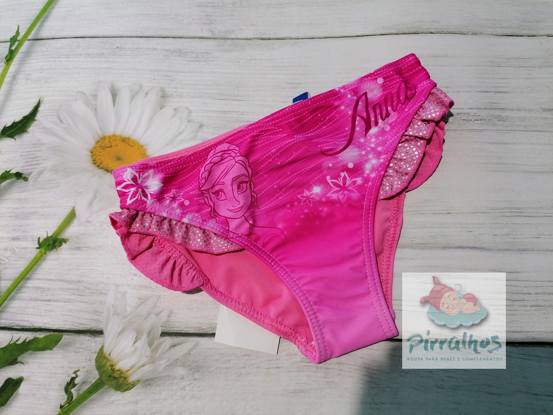 Cueca de banho infantil rosa com estampa da personagem Anna, babados e etiqueta Pirralhos sobre madeira branca com flores