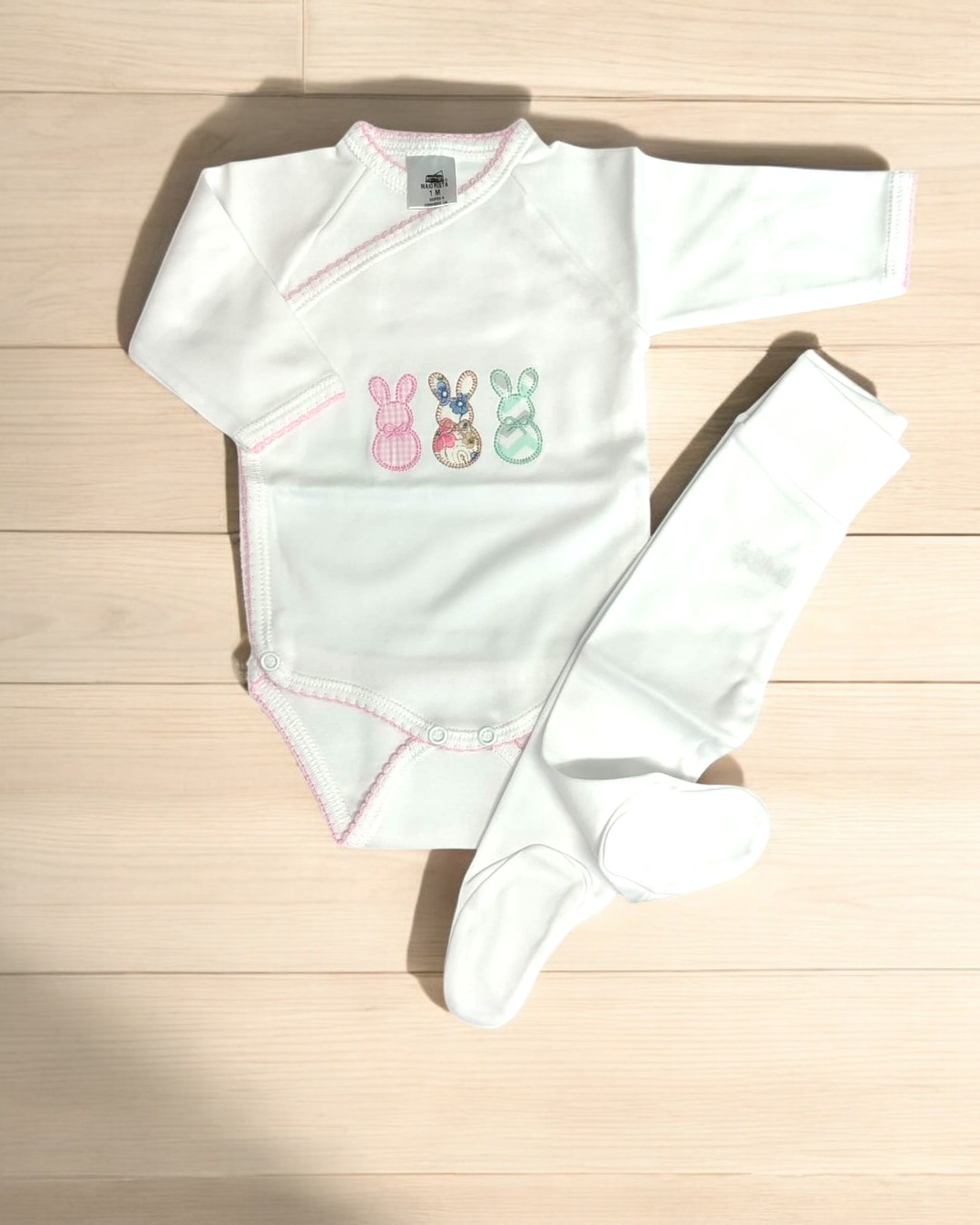 Conjunto de body branco com três coelhinhos bordados e meias brancas, em chão de madeira clara.