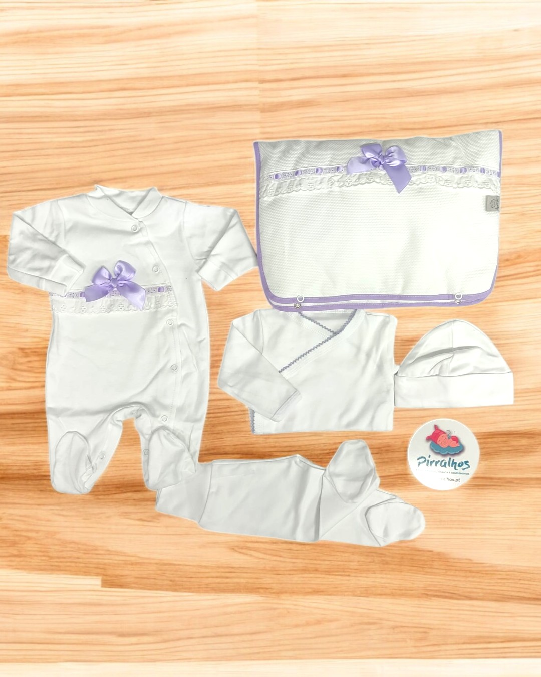 Conjunto de roupa de bebé branca com laço lilás e manta, em superfície de madeira