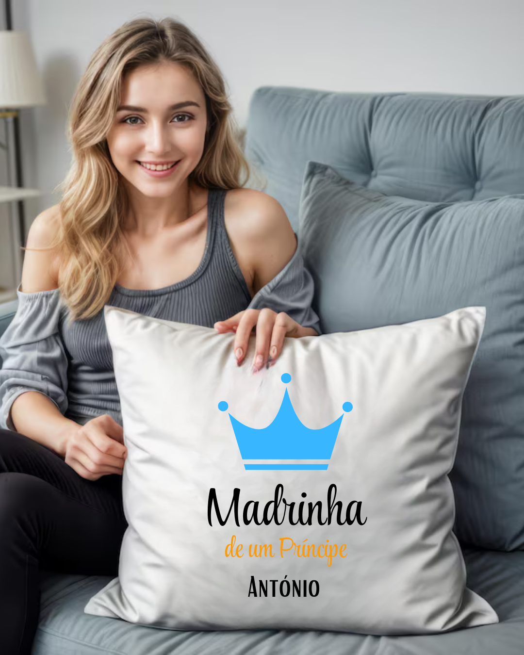 Almofada branca com coroa azul e texto 'Madrinha de um Príncipe ANTÓNIO' numa sala com sofá cinza