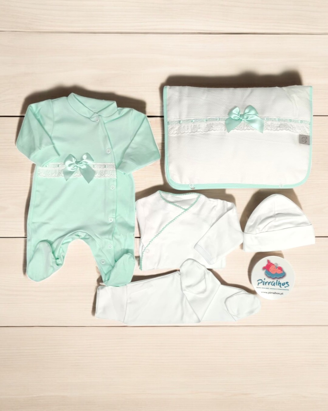 Conjunto de roupa de bebé verde menta e branco com laço e renda sobre piso claro.