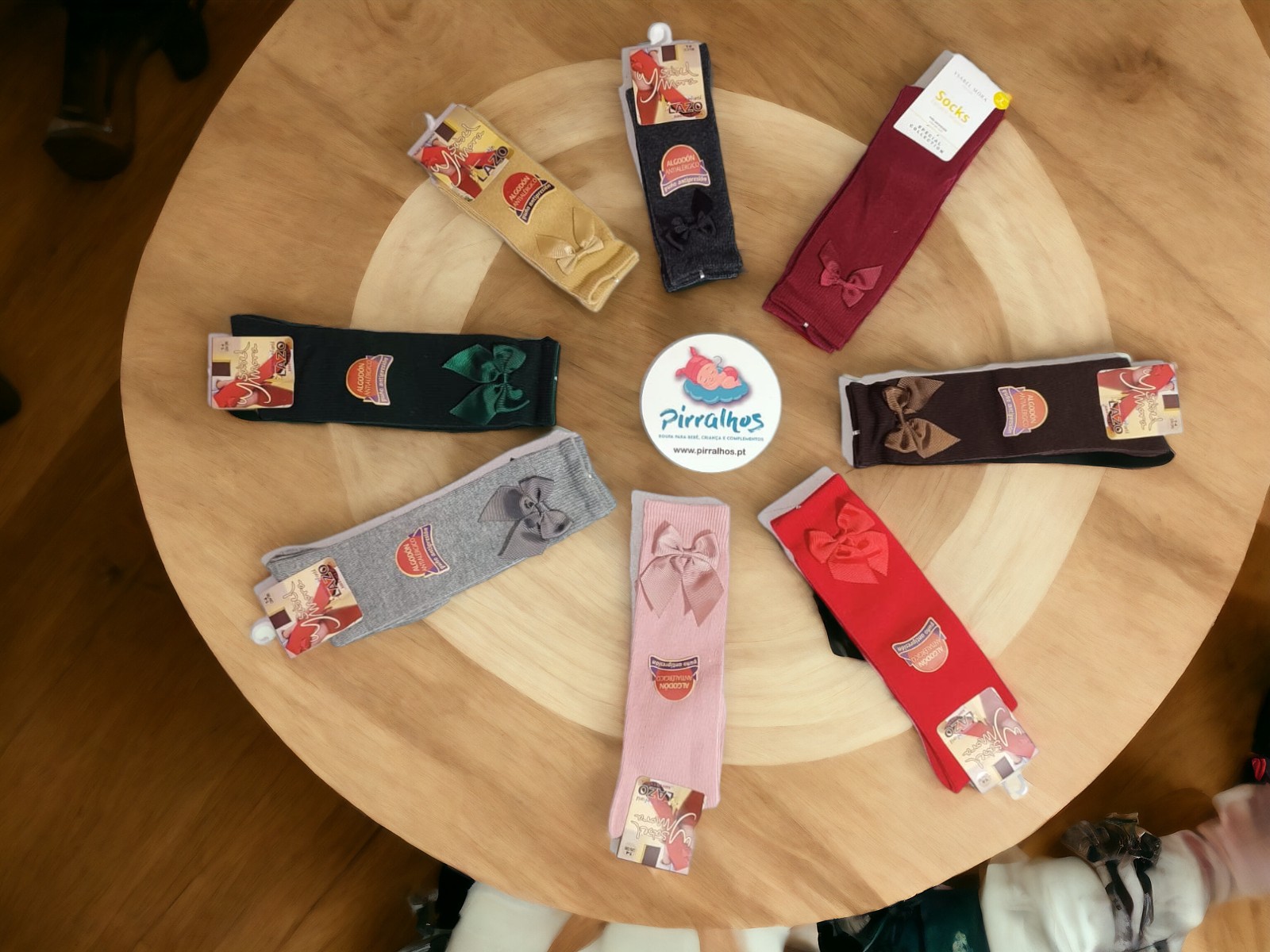 Oito pares de meias coloridas com laços dispostas em círculo sobre mesa de madeira e etiqueta redonda no centro.