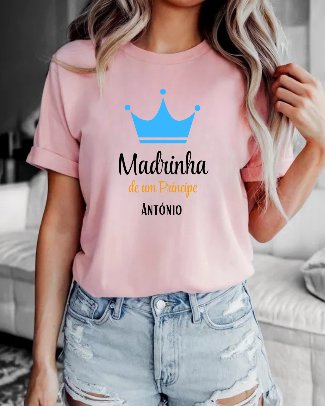 T-shirt rosa com coroa azul e texto 'Madrinha de um Príncipe ANTÓNIO', calções de ganga rasgados, ambiente interior