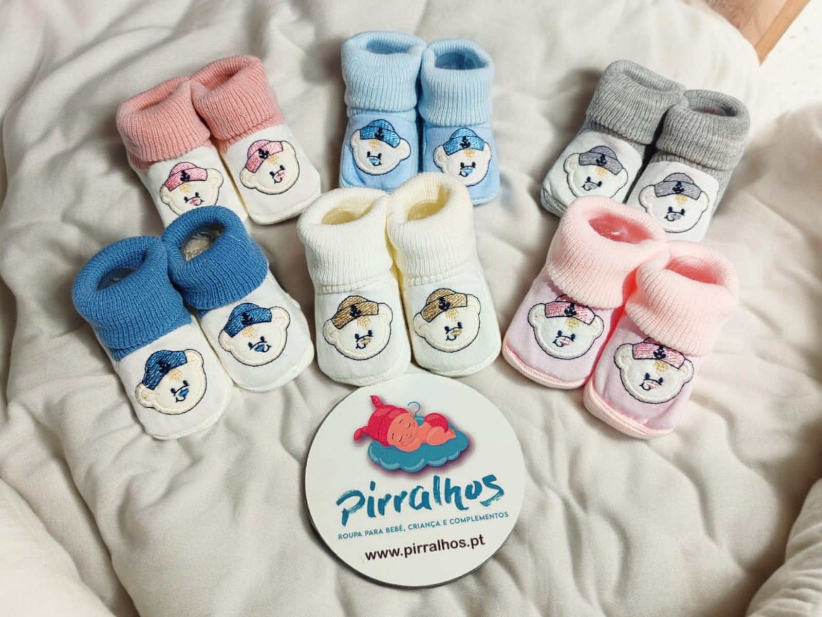 Seis pares de botins de bebé coloridos com cabeça de animal bordada, dispostos sobre tecido branco com cartão da marca Pirralhos.