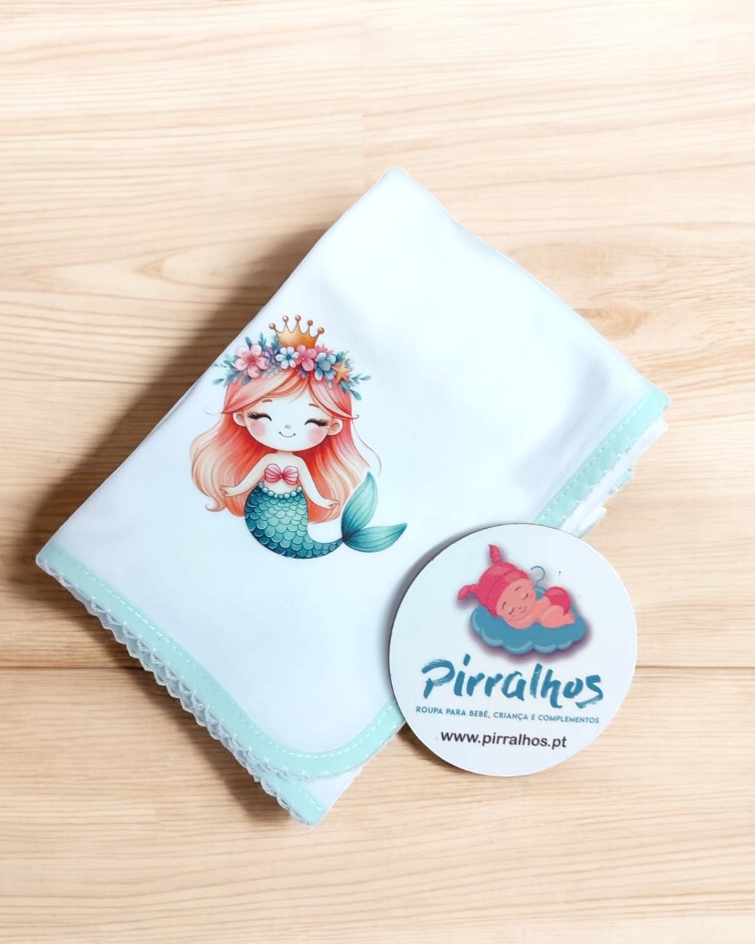 Manta branca com sereia estampada e cartão da marca Pirralhos num fundo de madeira clara