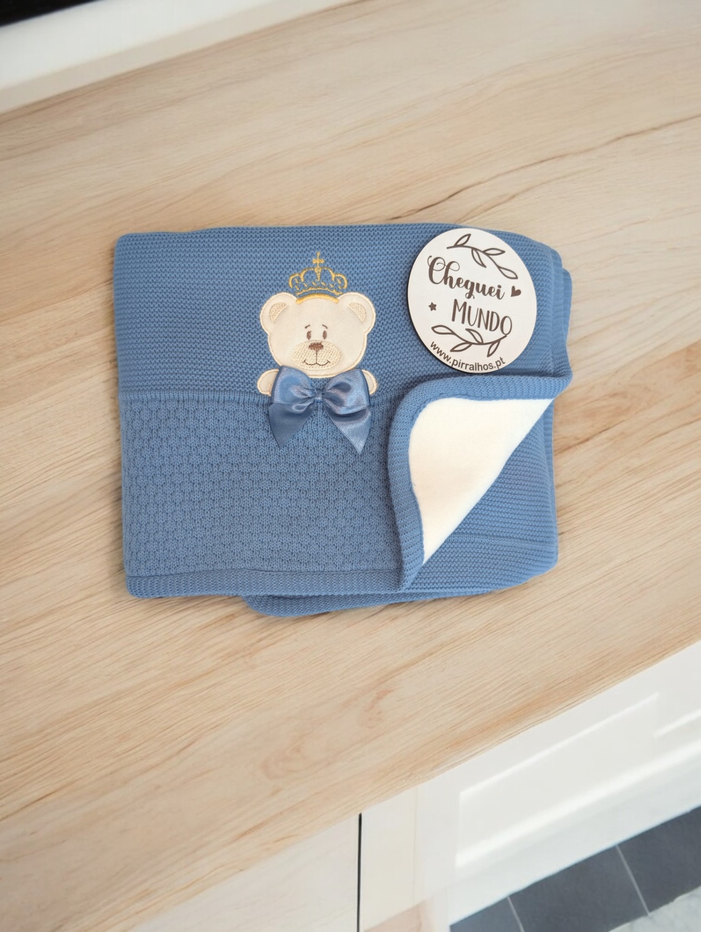 Manta azul de tricô com ursinho e etiqueta 'Cheguei ao MUNDO' sobre superfície de madeira