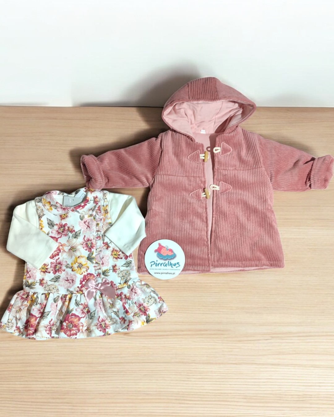 Conjunto de vestido florido e casaco rosa para bebé sobre mesa de madeira.