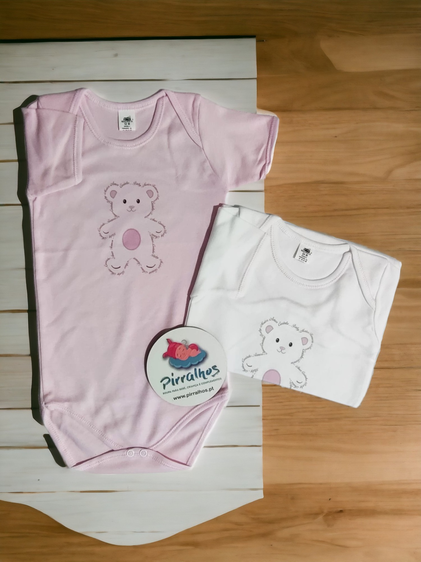 Dois bodies infantis, rosa e branco, com estampa de ursinho e selo da marca Pirralhos