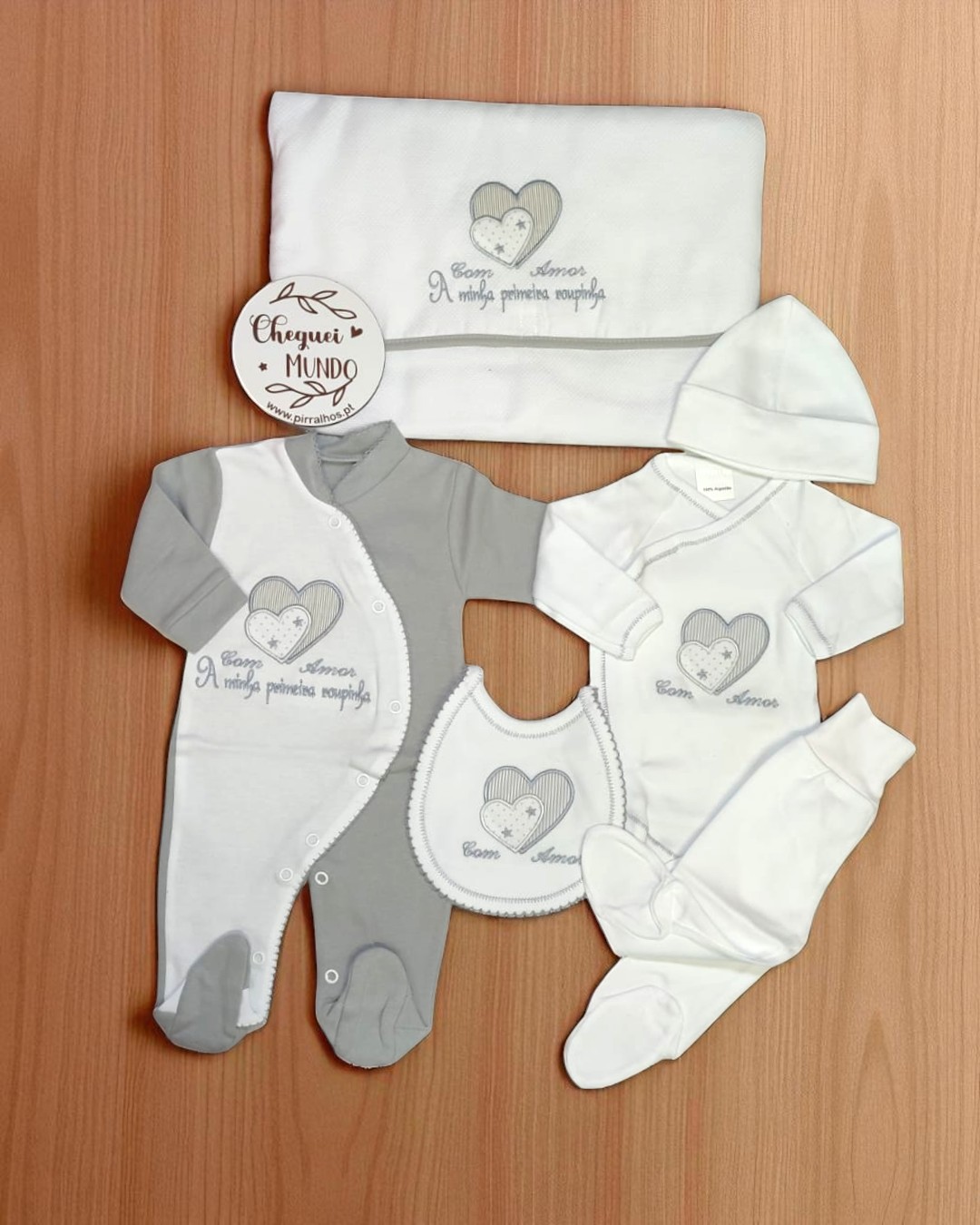 Conjunto de roupa branca e cinzenta para bebé com mensagens bordadas, com vários acessórios e placa de madeira decorativa.