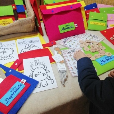 Material educativo colorido em mesa com pastas de feltro e cartões ilustrativos em mirandês.