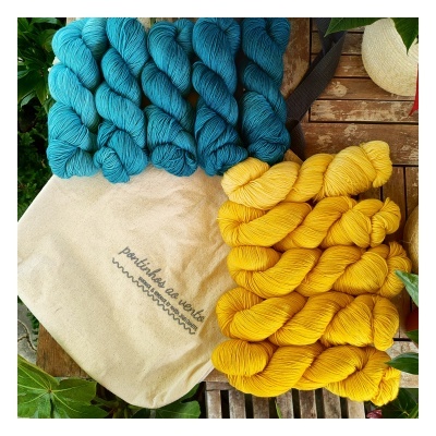 Novelos de lã azul e amarelo com saco de pano 'pontinho ao vento' sobre superfície de madeira