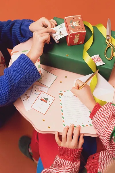 Crianças escrevendo cartões e etiquetas de Natal com material de embrulho festivo.