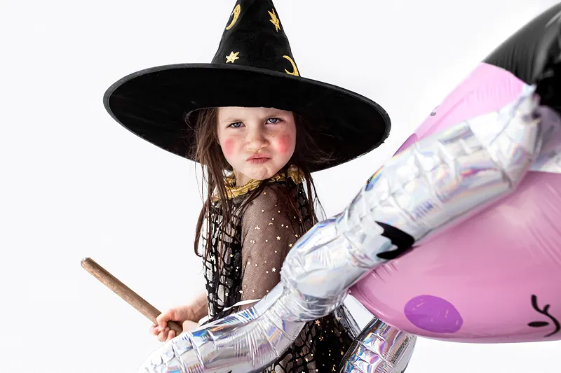 Criança vestida de bruxa com chapéu preto e varinha segurando balão inflável colorido