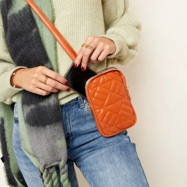 Bolsa pequena laranja matelassê com telemóvel sendo colocado dentro.