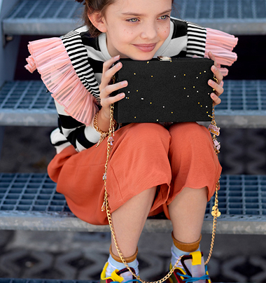 Mala preta com estrelas amarelas e corrente dourada, segurada por rapariga sentada em escadas metálicas