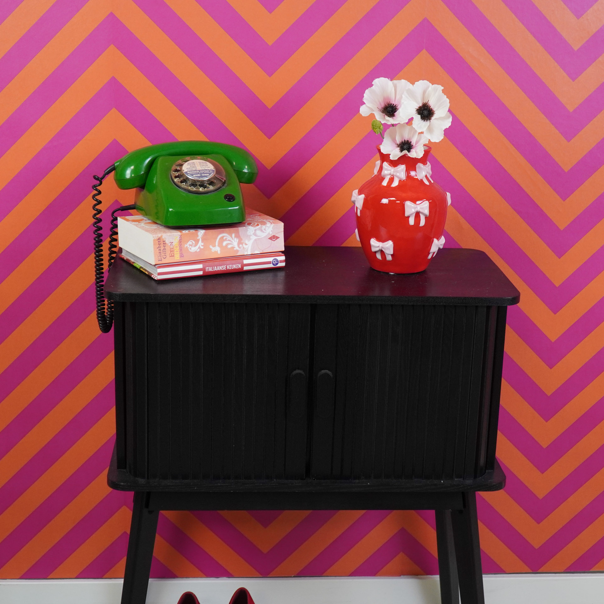Aparador preto com telefone verde, livros e vaso vermelho com flores, parede laranja e rosa ziguezague.