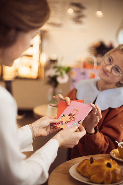 Pessoa entrega saco rosa decorado com chaveiro dourado em forma de animal a outra pessoa, numa mesa com doce em forma de coelho e chá