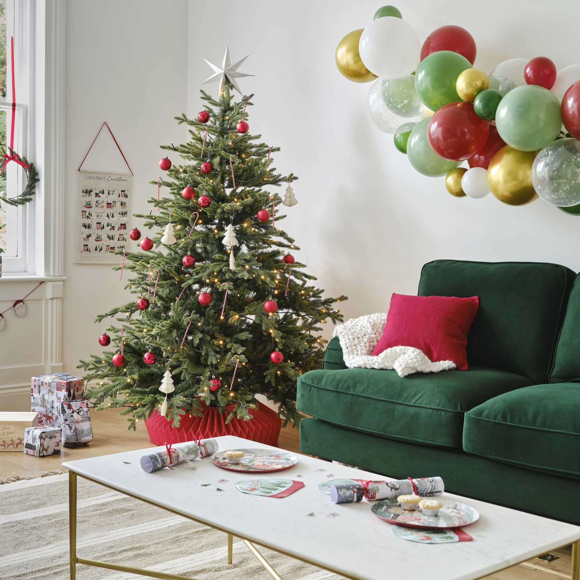 Árvore de Natal decorada com bolas vermelhas e enfeites brancos ao lado de sofá verde escuro e mesa branca com decorações
