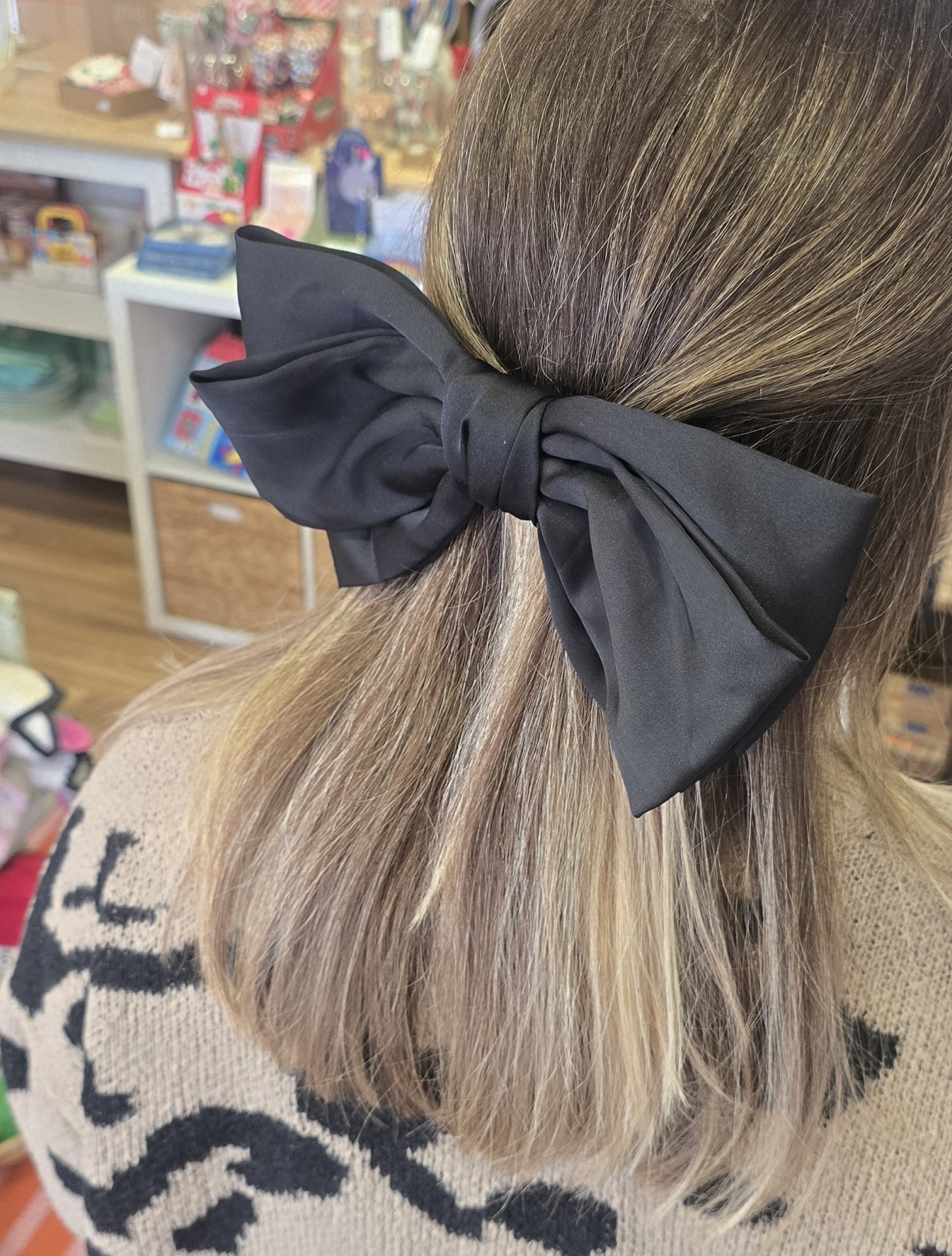 Laço preto grande no cabelo castanho claro de pessoa, prateleiras com objetos ao fundo.