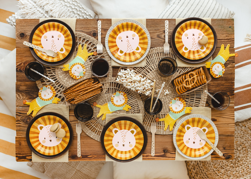 Mesa de madeira posta para festa infantil com pratos e decoração de leões e snacks variados
