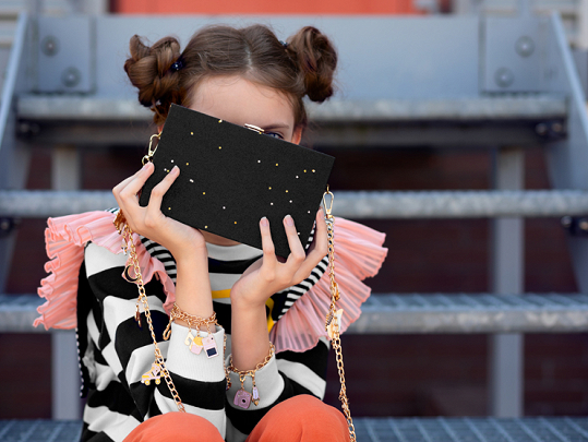 Carteira preta com pontos dourados e correia de corrente, segurada por pessoa com camisola às riscas e folhos cor de rosa