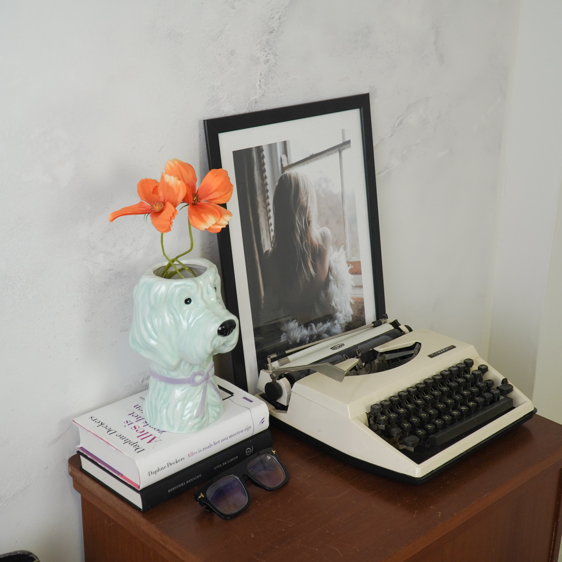 Máquina de escrever, fotografia, vaso decorativo com flores, livros e óculos escuros sobre móvel de madeira