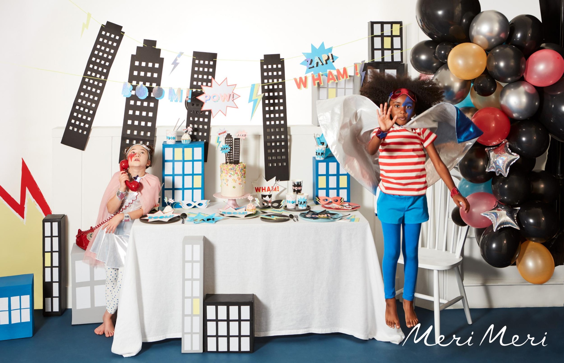 Crianças em festa temática com decoração de prédios e balões coloridos.