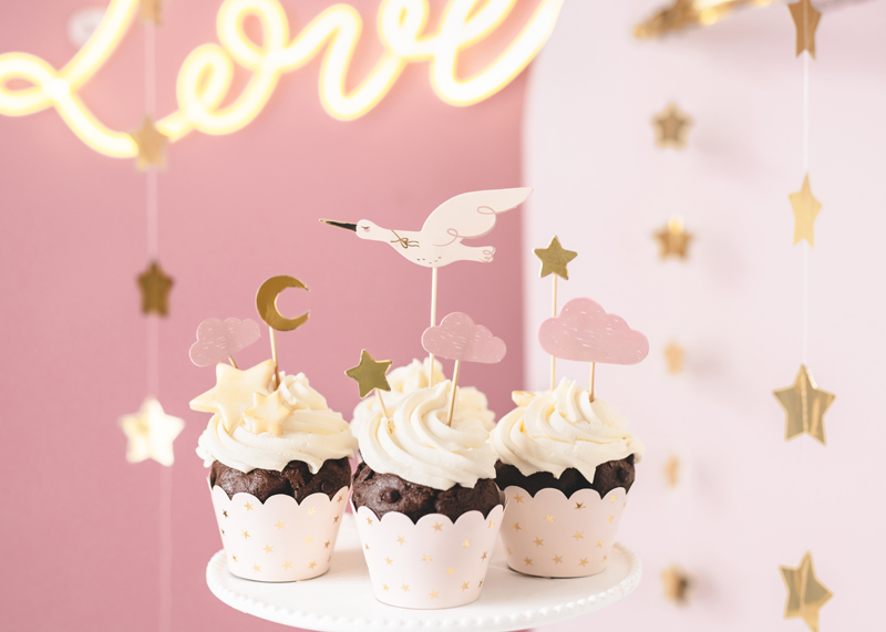 Três cupcakes decorados com coberturas brancas e toppers temáticos num suporte branco, com fundo rosa e estrelas douradas pendentes.
