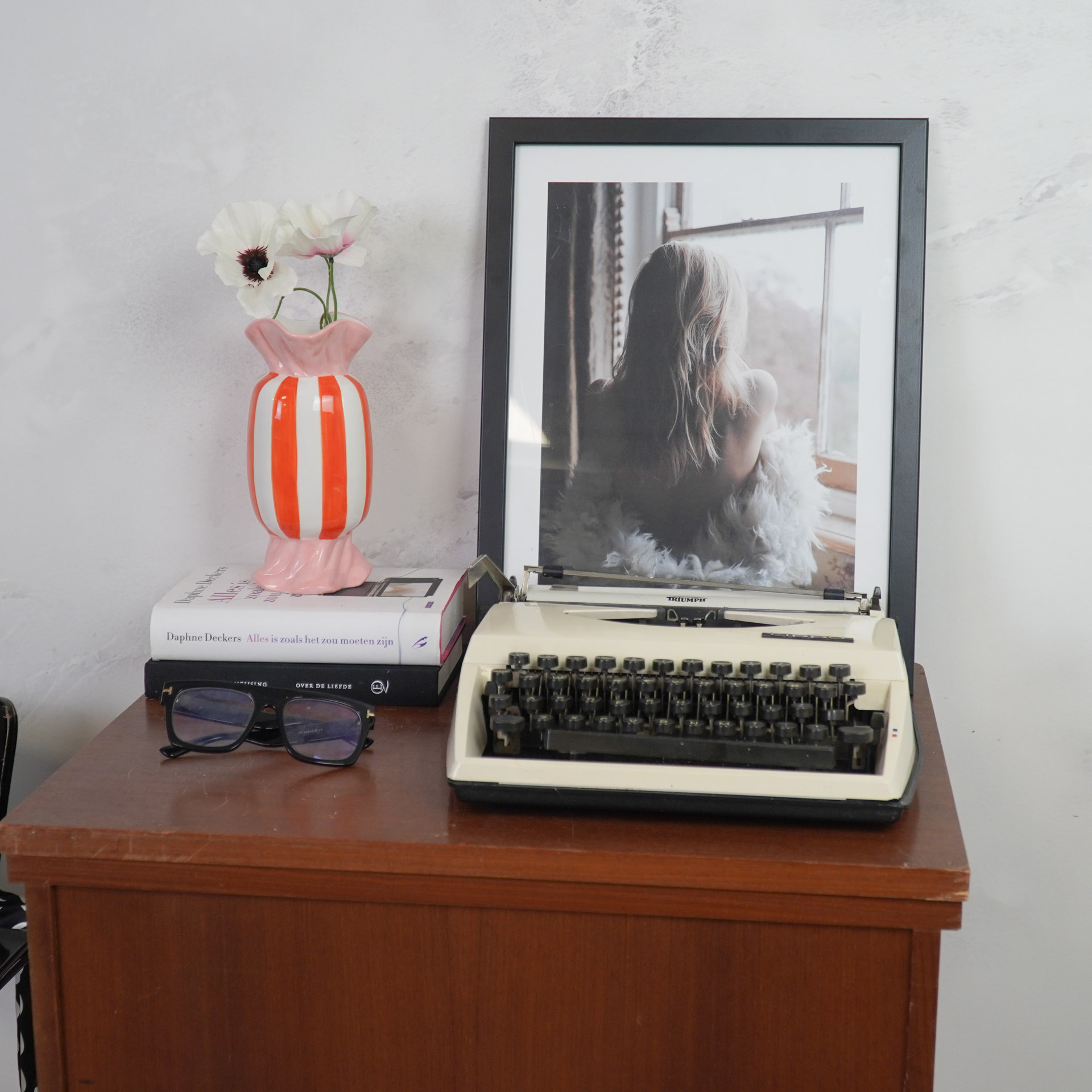 Secretária com máquina de escrever, fotografia, vaso com flores, livros e óculos.