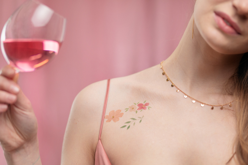 Colar dourado com pendentes e tatuagens florais num ombro, mulher com vestido rosa segurando copo de vinho rosé.