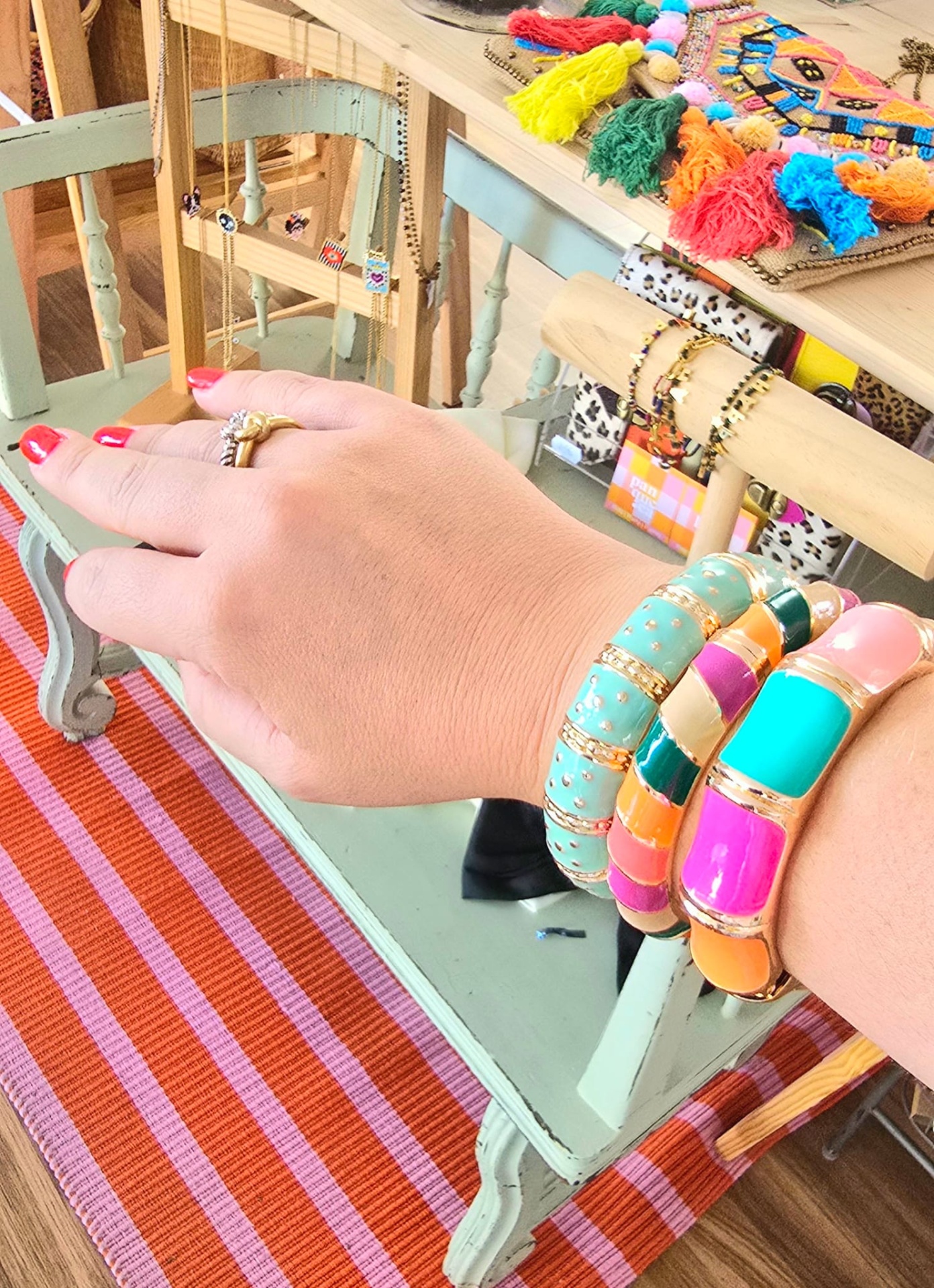 Braços com pulseiras coloridas e anel sobre banco verde claro, tapete listrado vermelho e acessórios variados ao fundo