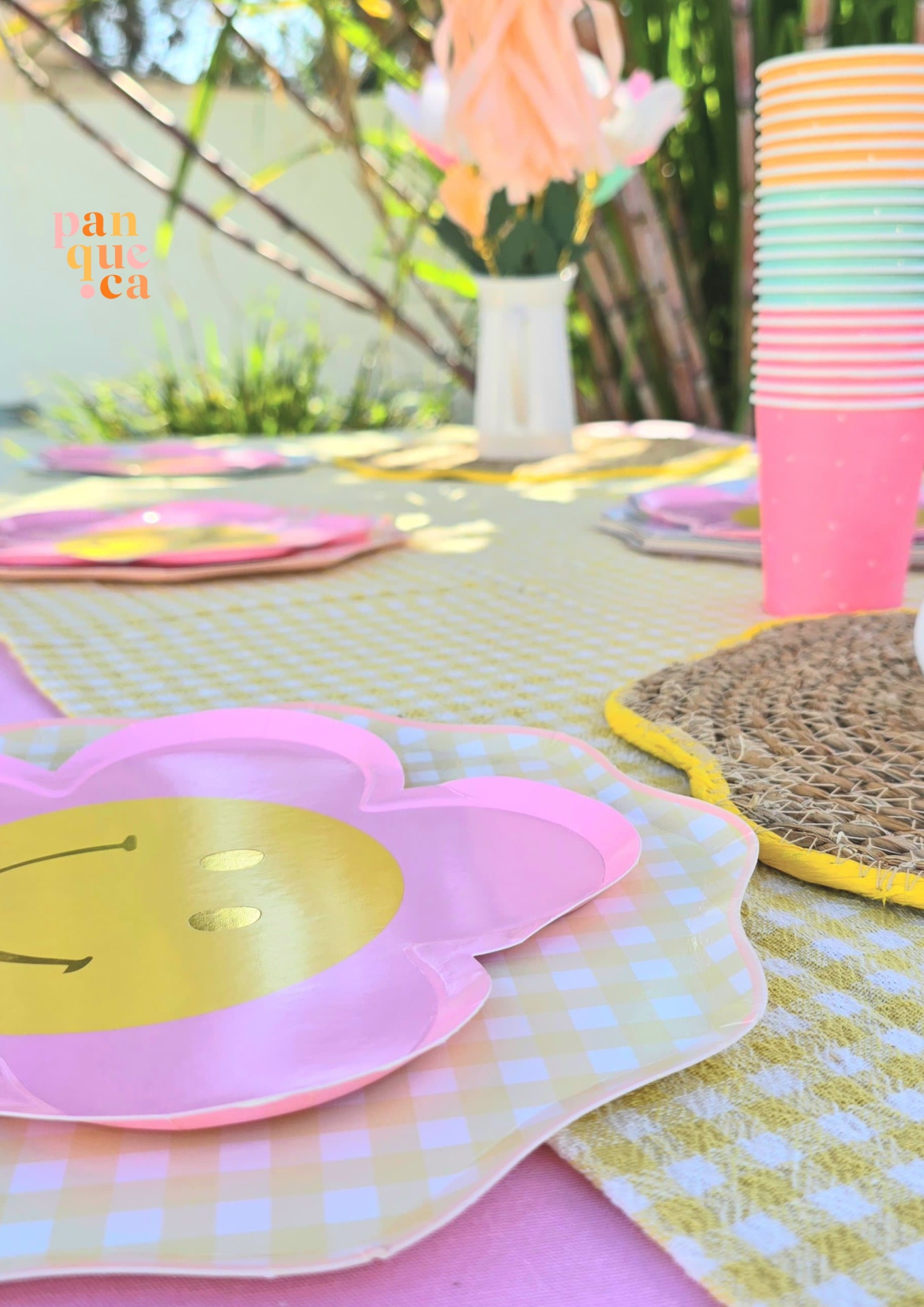 Mesa de festa com pratos de flores cor-de-rosa e bege, copos coloridos empilhados e arranjo floral em vaso branco