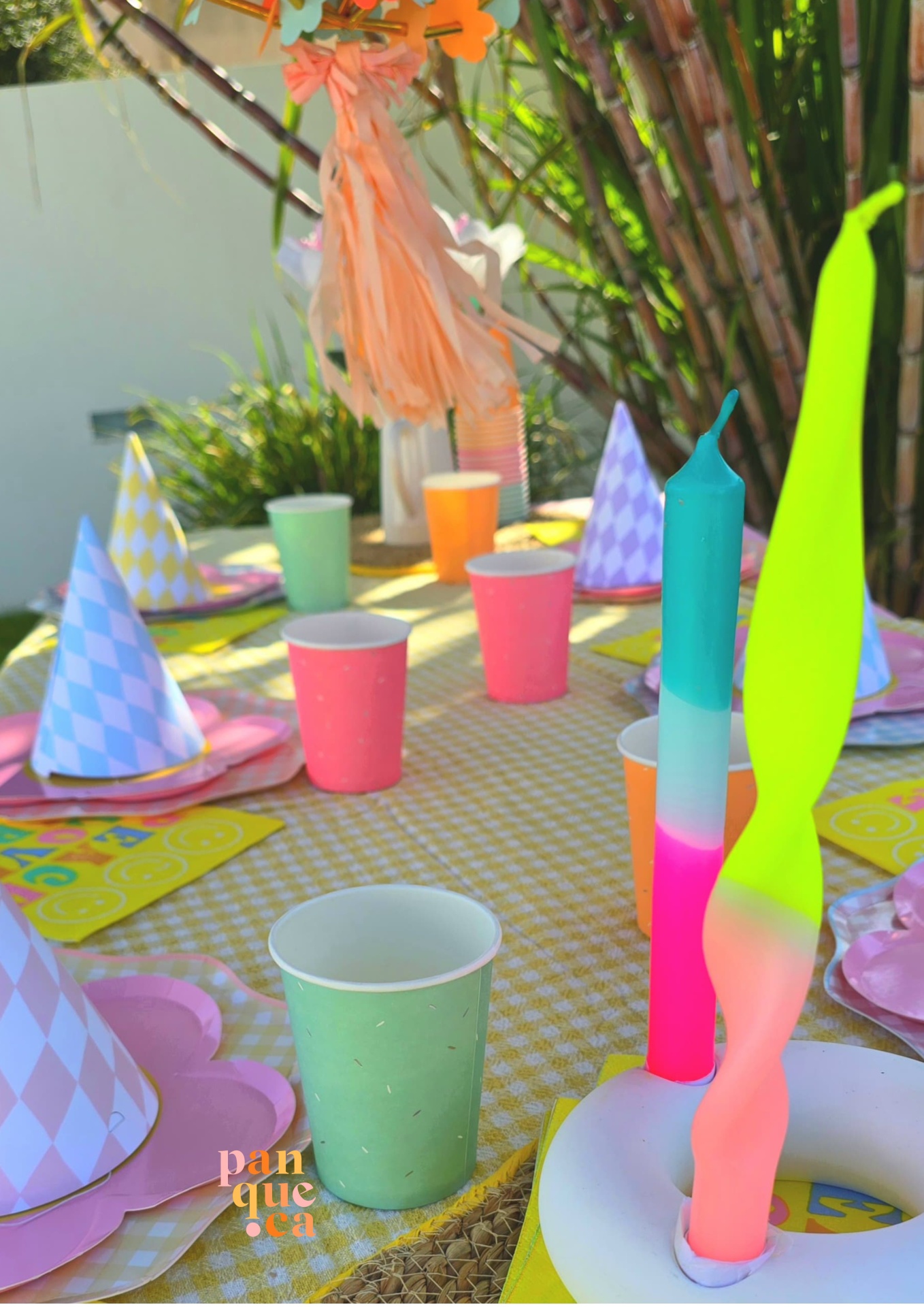 Mesa de festa decorada com toalha amarela xadrez, chapéus de cone lilás e branco, copos coloridos e velas torcidas coloridas