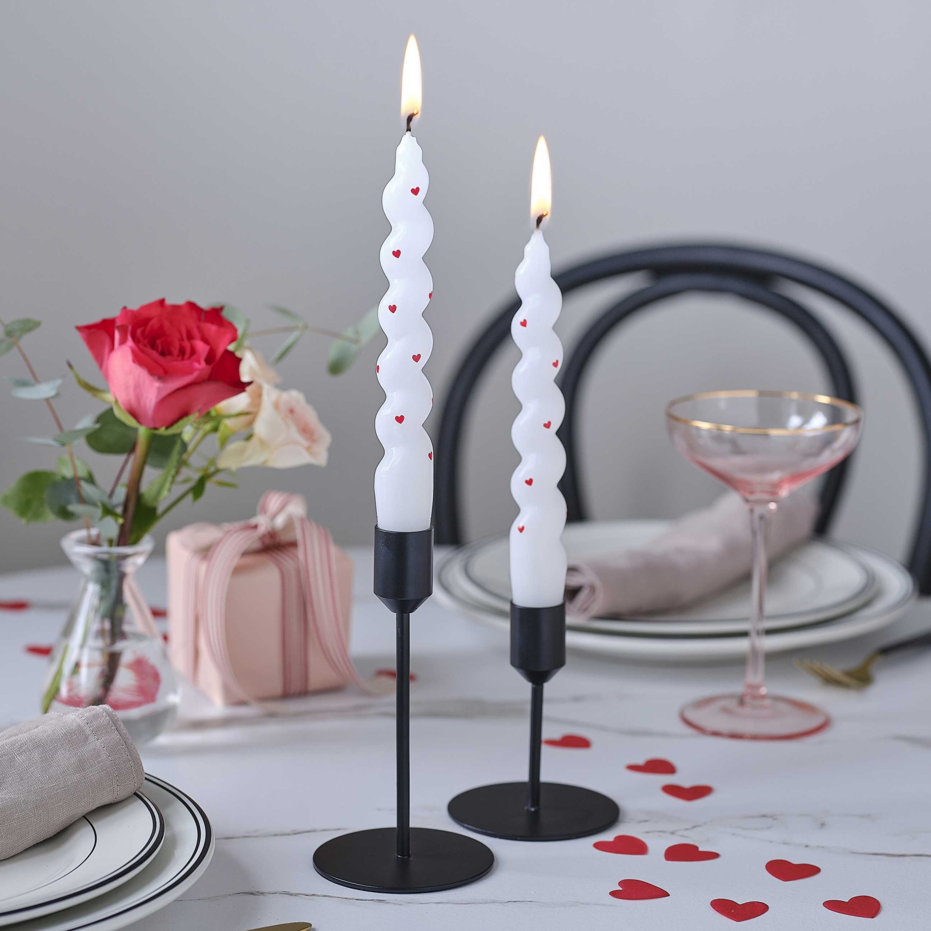 Velas brancas com corações vermelhos em castiçais pretos numa mesa decorada com flores e presente