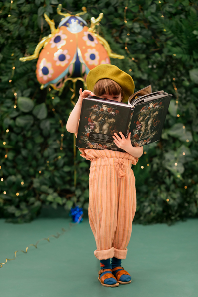 Criança com calças cor de pêssego e boina lendo livro diante de decoração com joaninha laranja