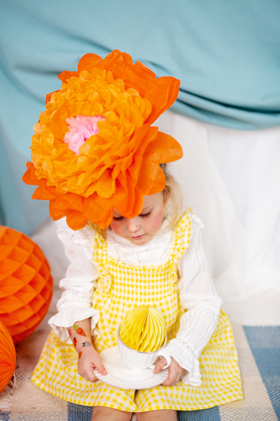 Criança com vestido amarelo e chapéu de flor laranja segurando chávena com ornamento de papel amarelo