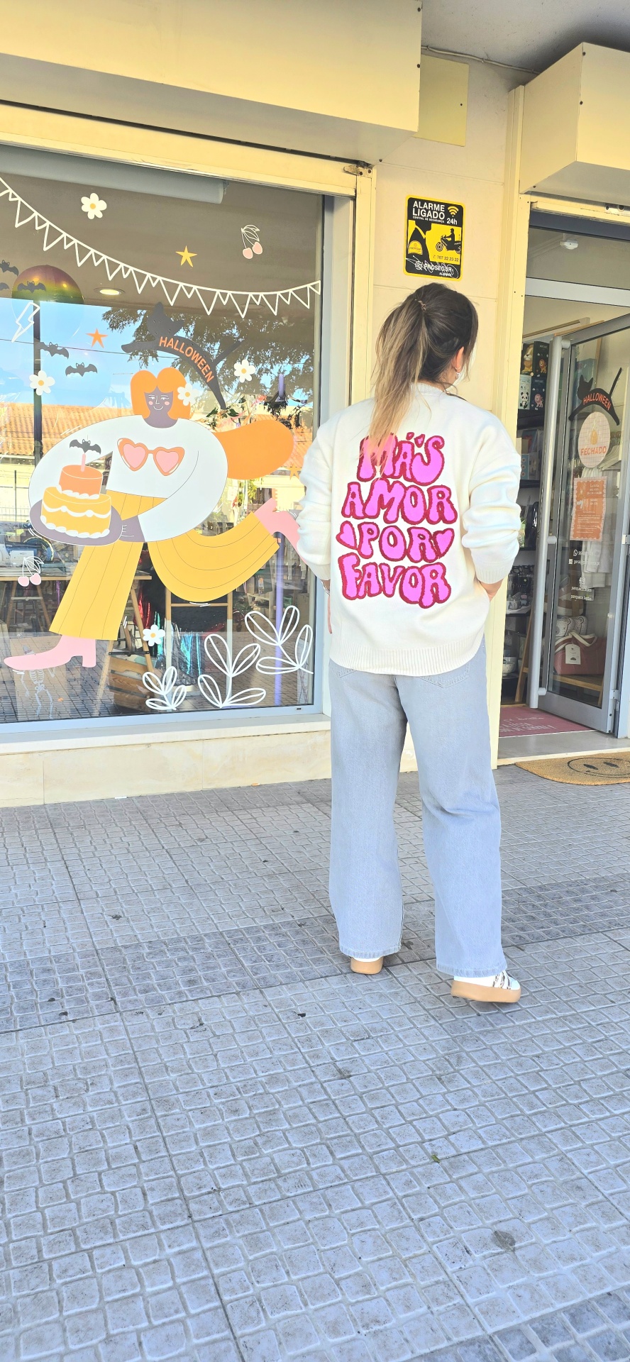 Pessoa de costas com casaco branco estampado rosa e calças de ganga em frente a loja com decoração de Halloween