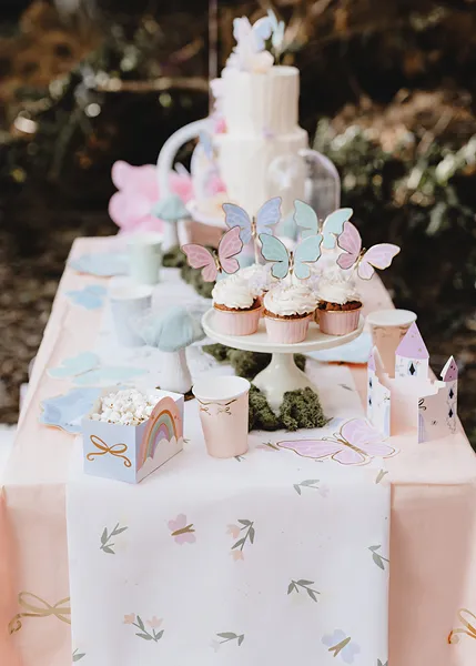 Mesa de festa decorada com cupcakes com borboletas, toalha branca com padrão, caixas decorativas e bolo branco