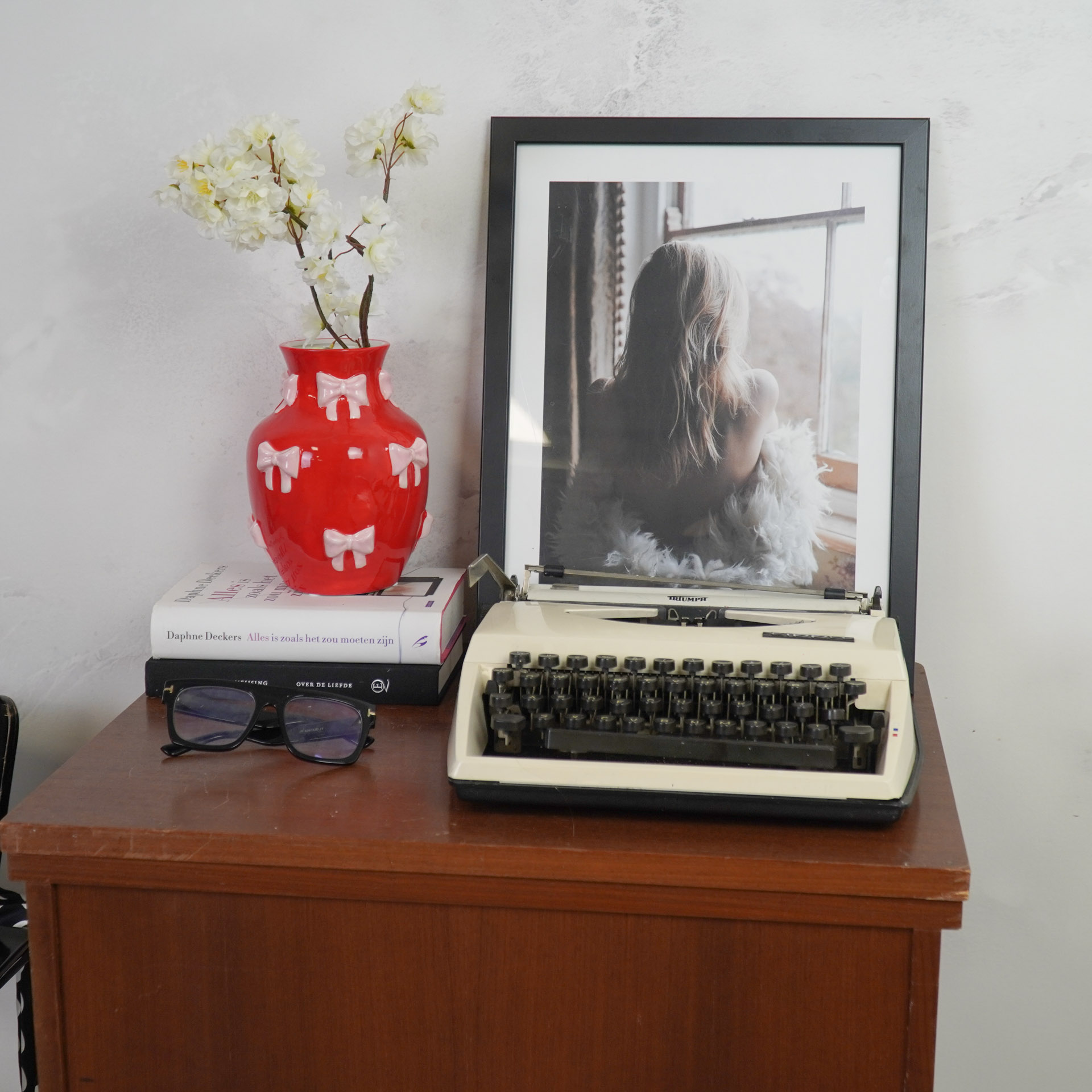 Mesa com máquina de escrever Triumph, vaso vermelho com flores, livros e óculos