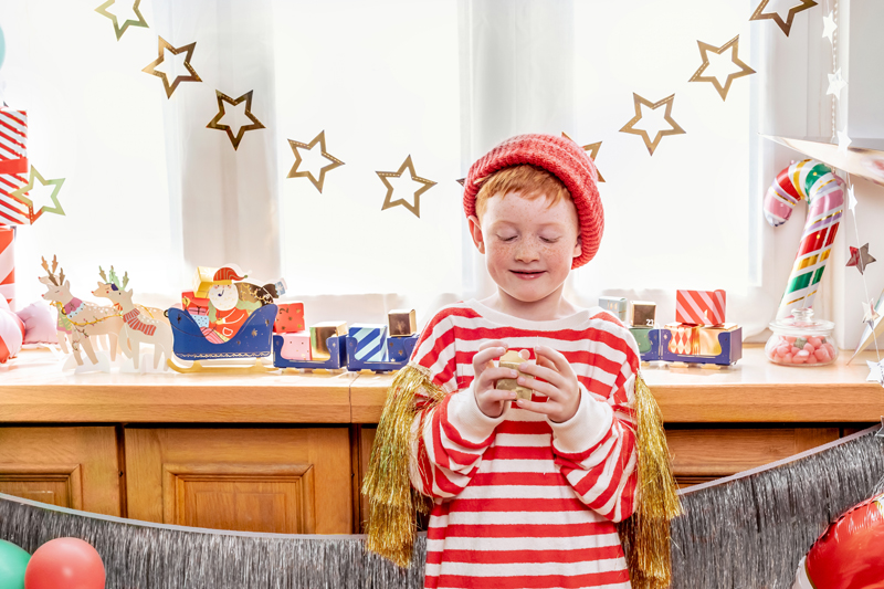 Criança com camisola às riscas e gorro vermelho segurando caneca em ambiente decorado de Natal