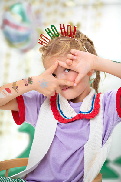 Criança com roupa colorida e fita de cabelo 'HO HO HO!' faz moldura com as mãos no rosto